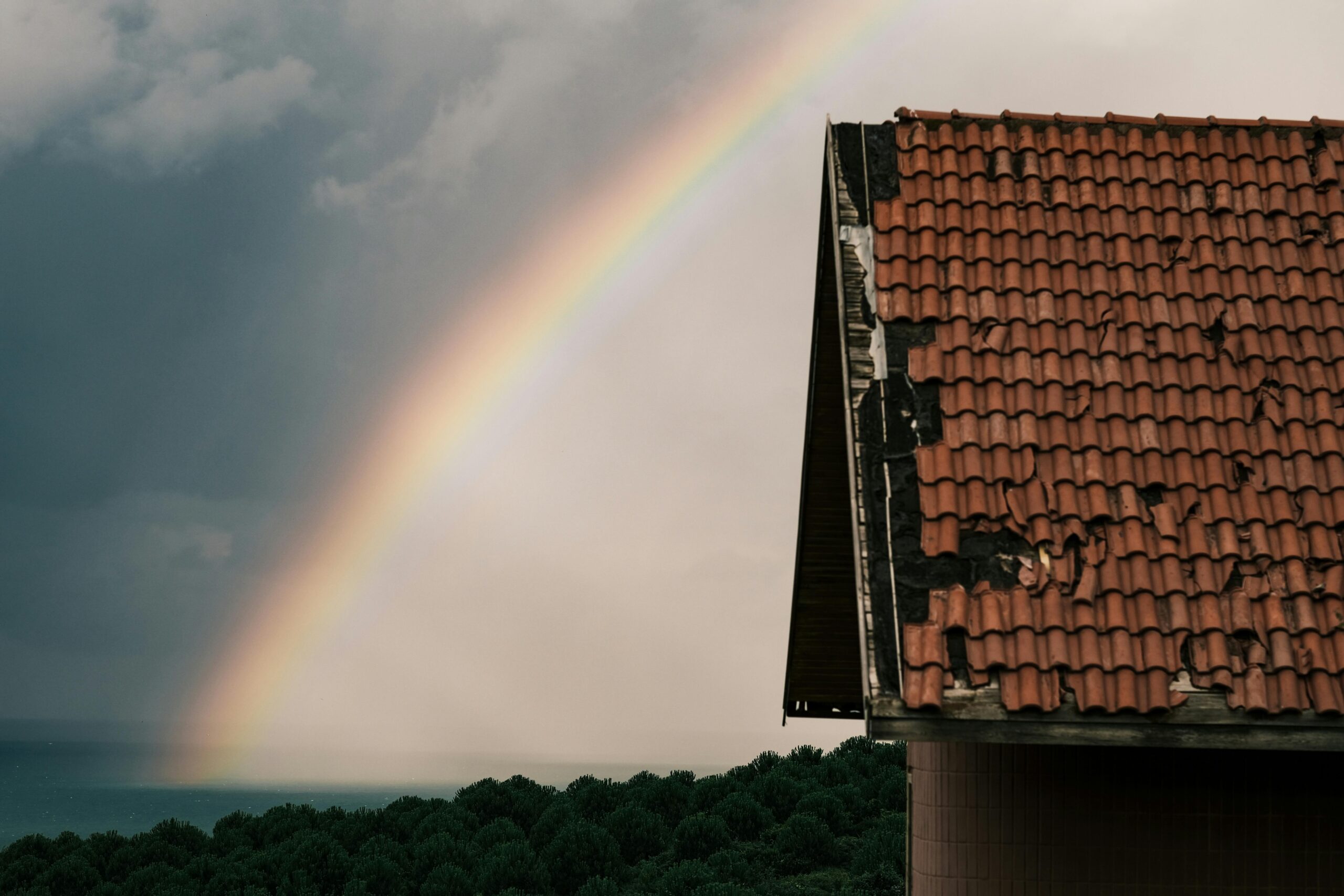photograph of shingles on a roof torn off after a storm. there are grey skies and a rainbow in the background